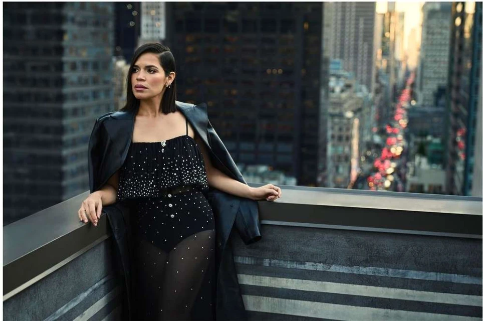 Adult female in a black embellished bodysuit and leather coat posing on a rooftop with a city skyline background and sunset light.