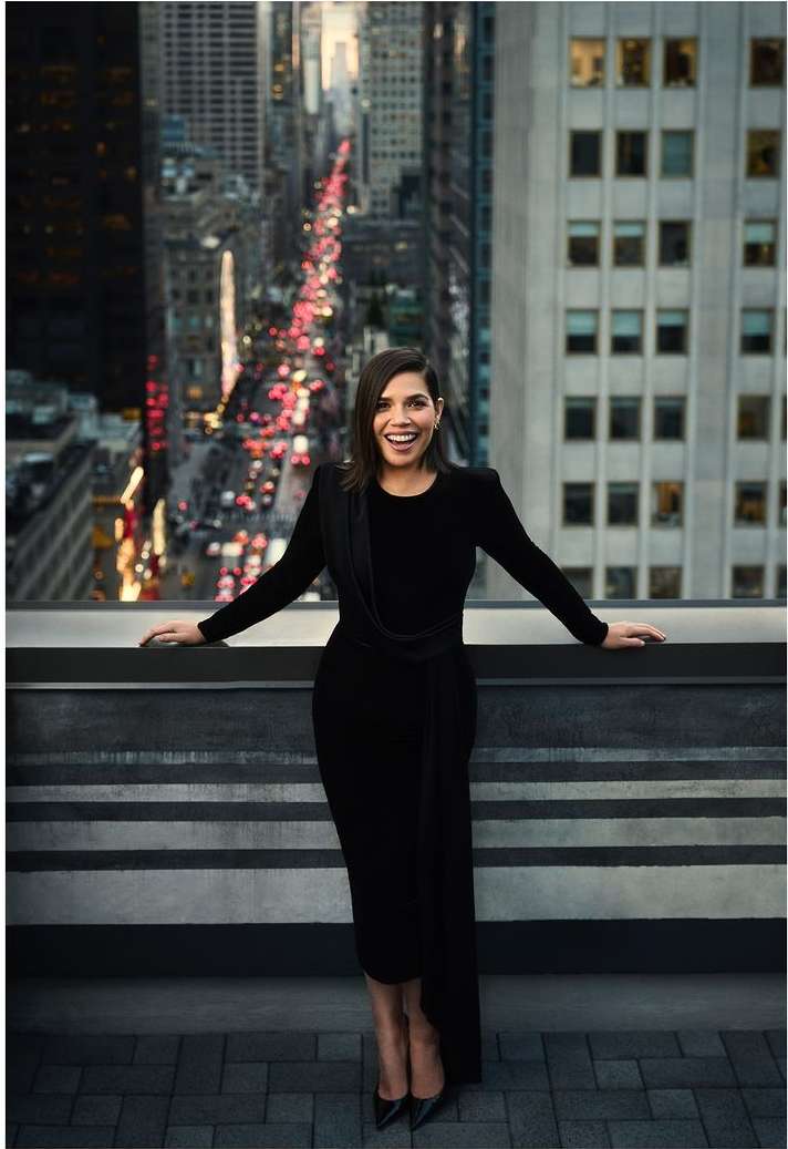 A woman in a black dress stands on a rooftop overlooking a city street with high-rise buildings, posing for a fashion editorial portrait.