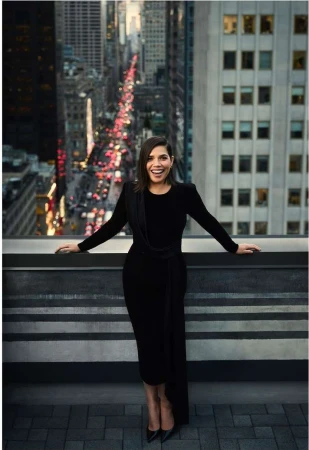 A woman in a black dress stands on a rooftop overlooking a city street with high-rise buildings, posing for a fashion editorial portrait.