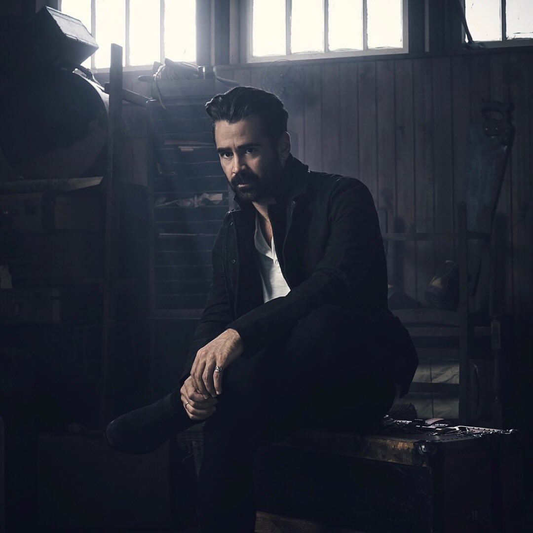 Portrait of Colin Farrell with a dark beard wearing a black jacket and white t-shirt sitting in an industrial interior.