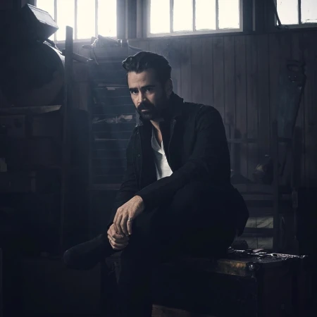 Portrait of Colin Farrell with a dark beard wearing a black jacket and white t-shirt sitting in an industrial interior.