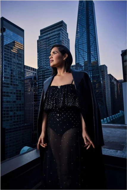Woman in black gown poses in an urban setting. Cityscape with tall buildings rises behind her in an eye-level portrait shot.