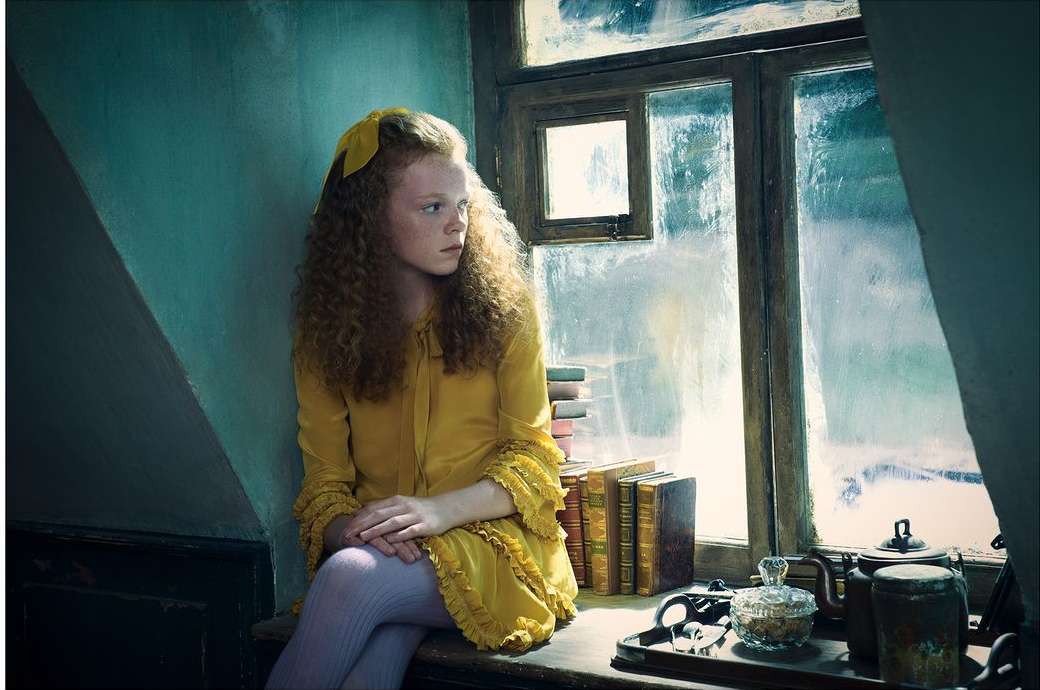 Young girl with curly hair in yellow dress and bow, sitting near window with books and teaware, in an indoor setting.