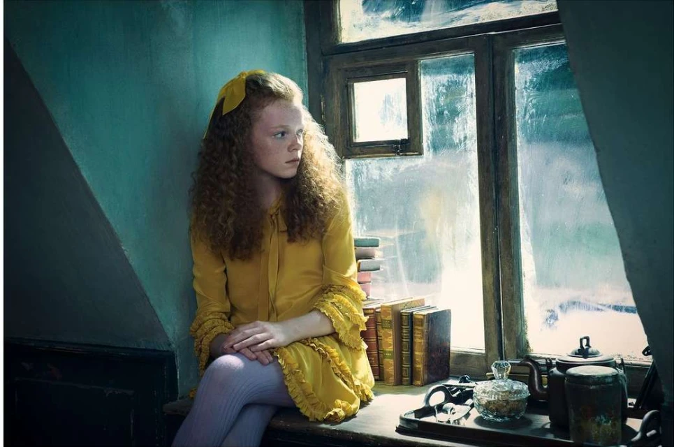 Young girl with curly hair in yellow dress and bow, sitting near window with books and teaware, in an indoor setting.