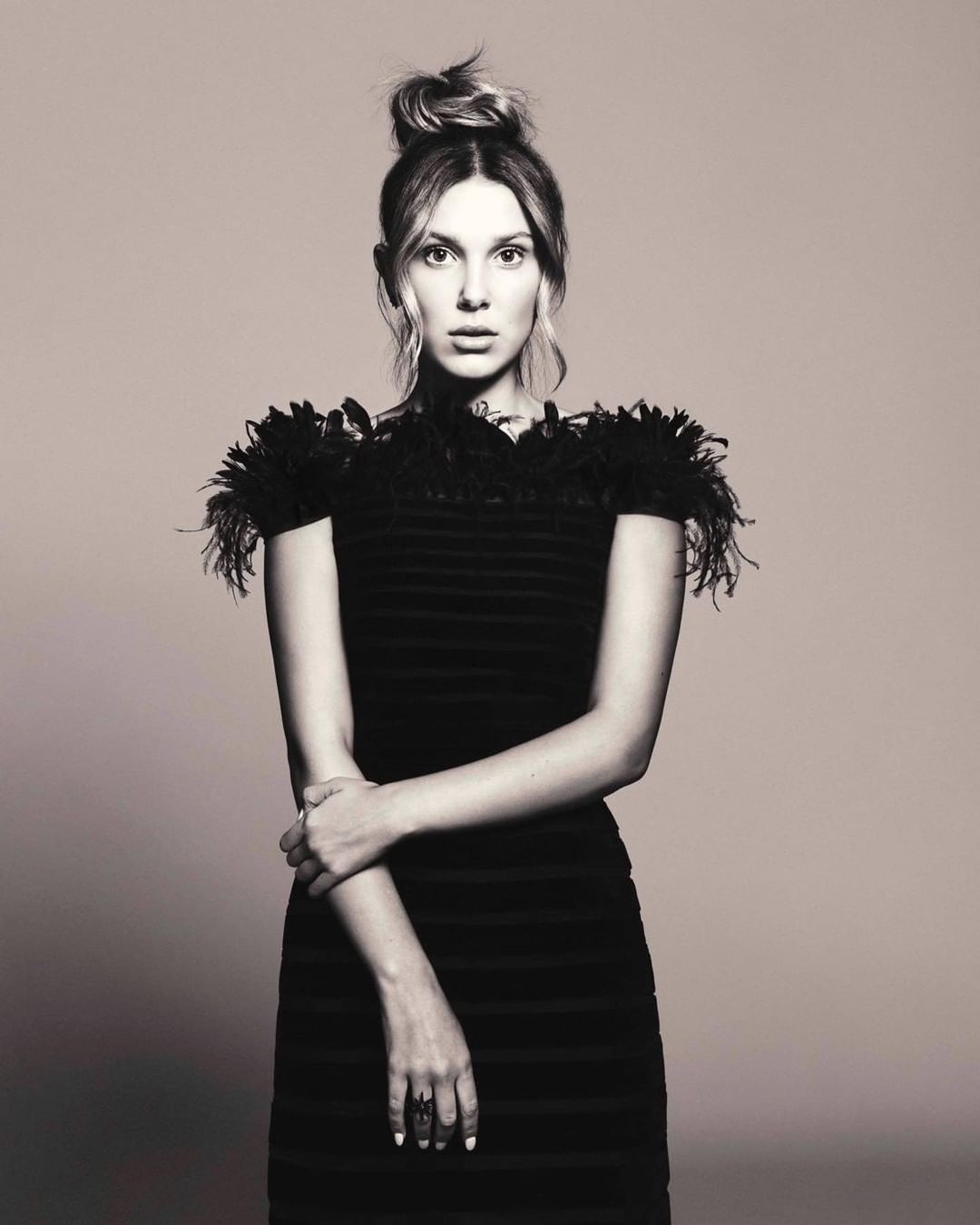 Portrait of a young woman in a black dress with feather details, posing with arms crossed in a studio with a neutral background.