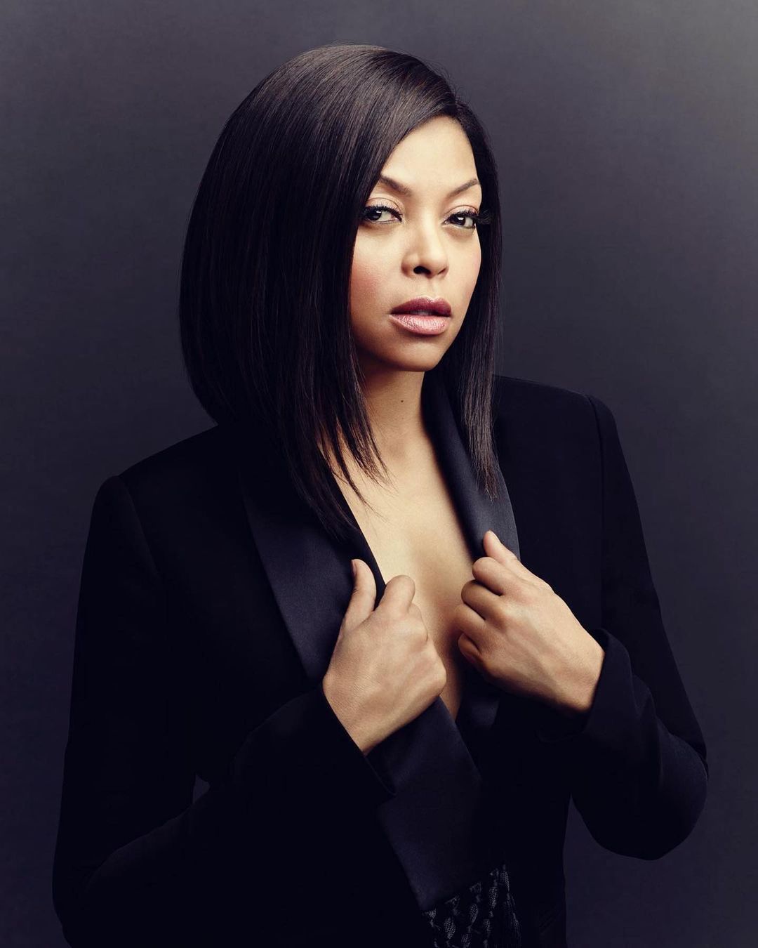 Glamour shot of an African American woman in a black blazer with sleek black hair against a dark grey backdrop in a studio.