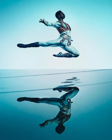 Male ballet dancer in ornate costume leaps above reflective water surface against a light blue gradient backdrop in a studio shot.