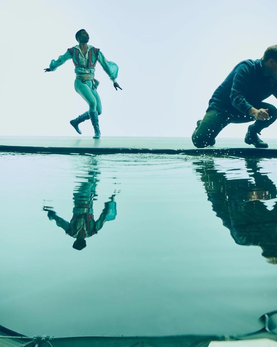 Male ballet dancer in a light blue costume poses gracefully above a pool of water with a second man in jeans crouched in the background.