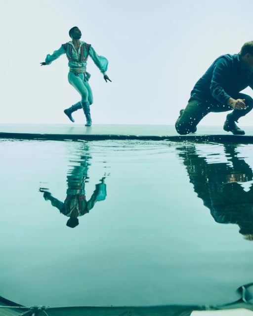 Male ballet dancer in a light blue costume poses gracefully above a pool of water with a second man in jeans crouched in the background.
