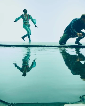 Male ballet dancer in a light blue costume poses gracefully above a pool of water with a second man in jeans crouched in the background.