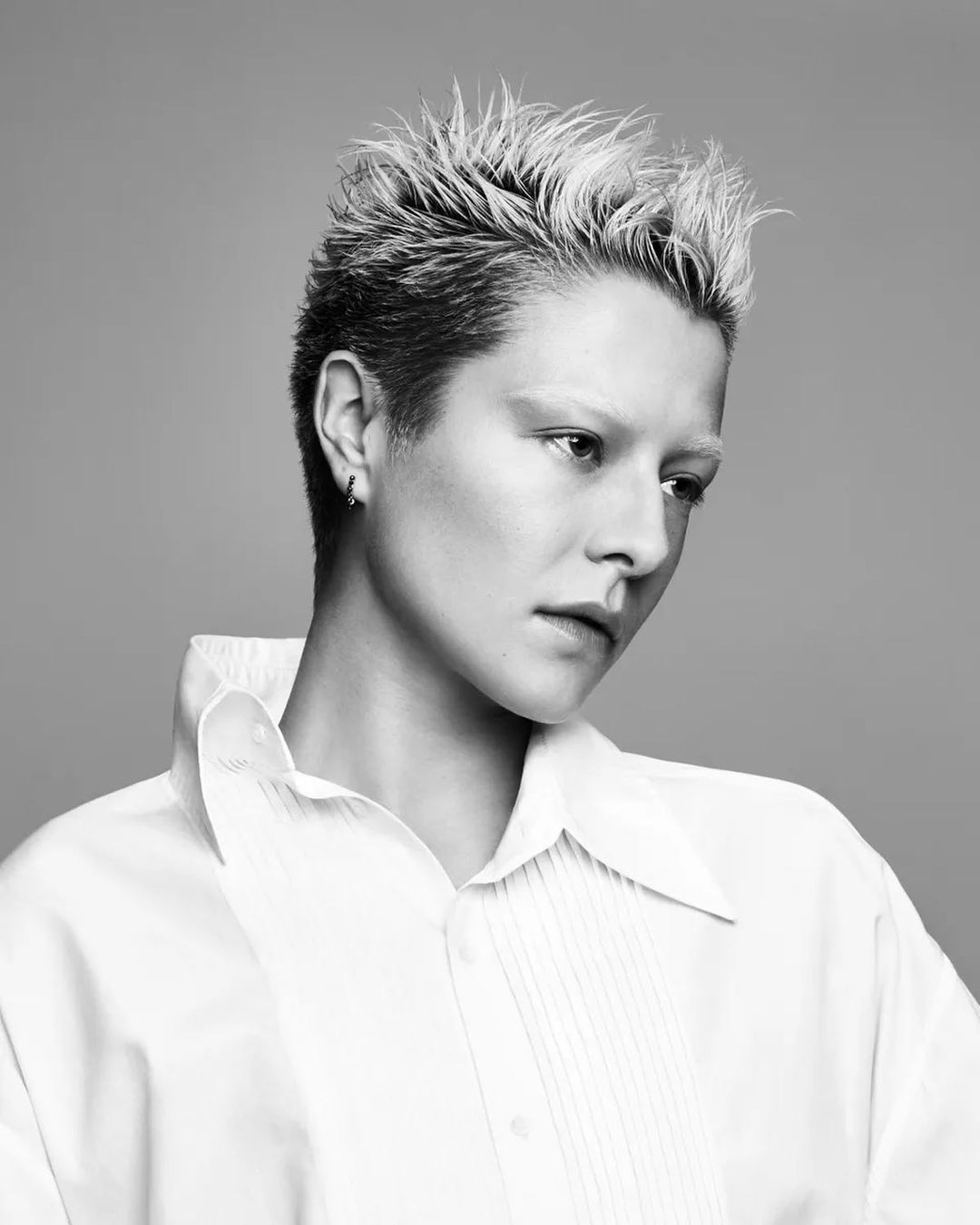 Monochrome portrait of a young adult with bleached spiky hair wearing a white collared shirt against a neutral gray backdrop.