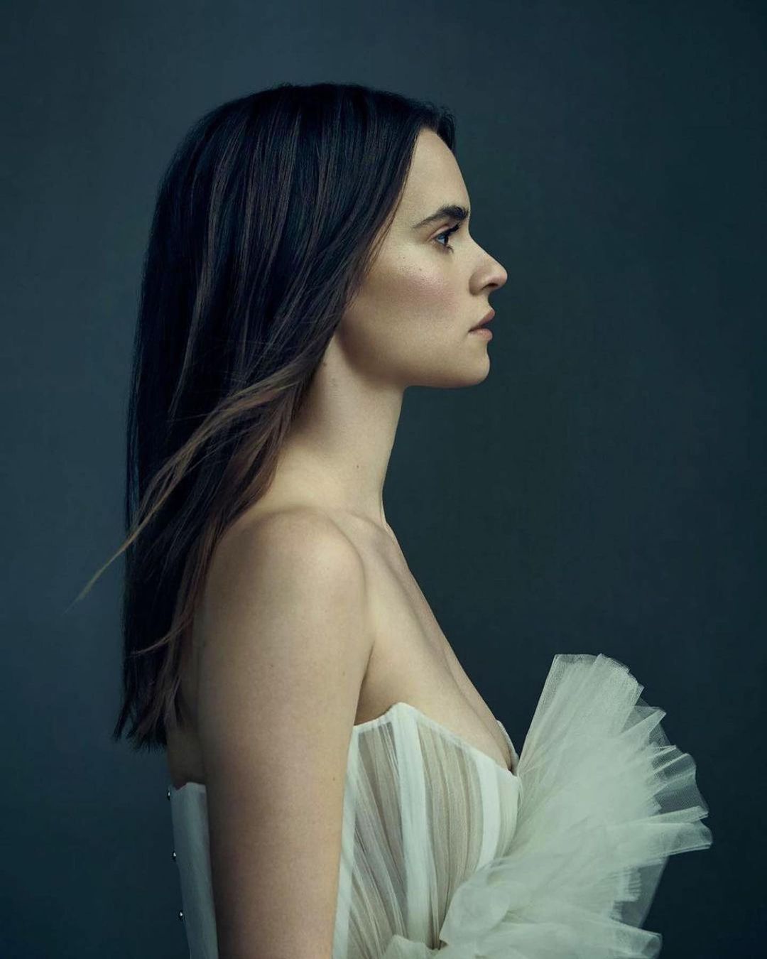 Portrait of a young woman in a strapless white dress against a solid blue-gray background, medium shot, studio lighting, side profile.