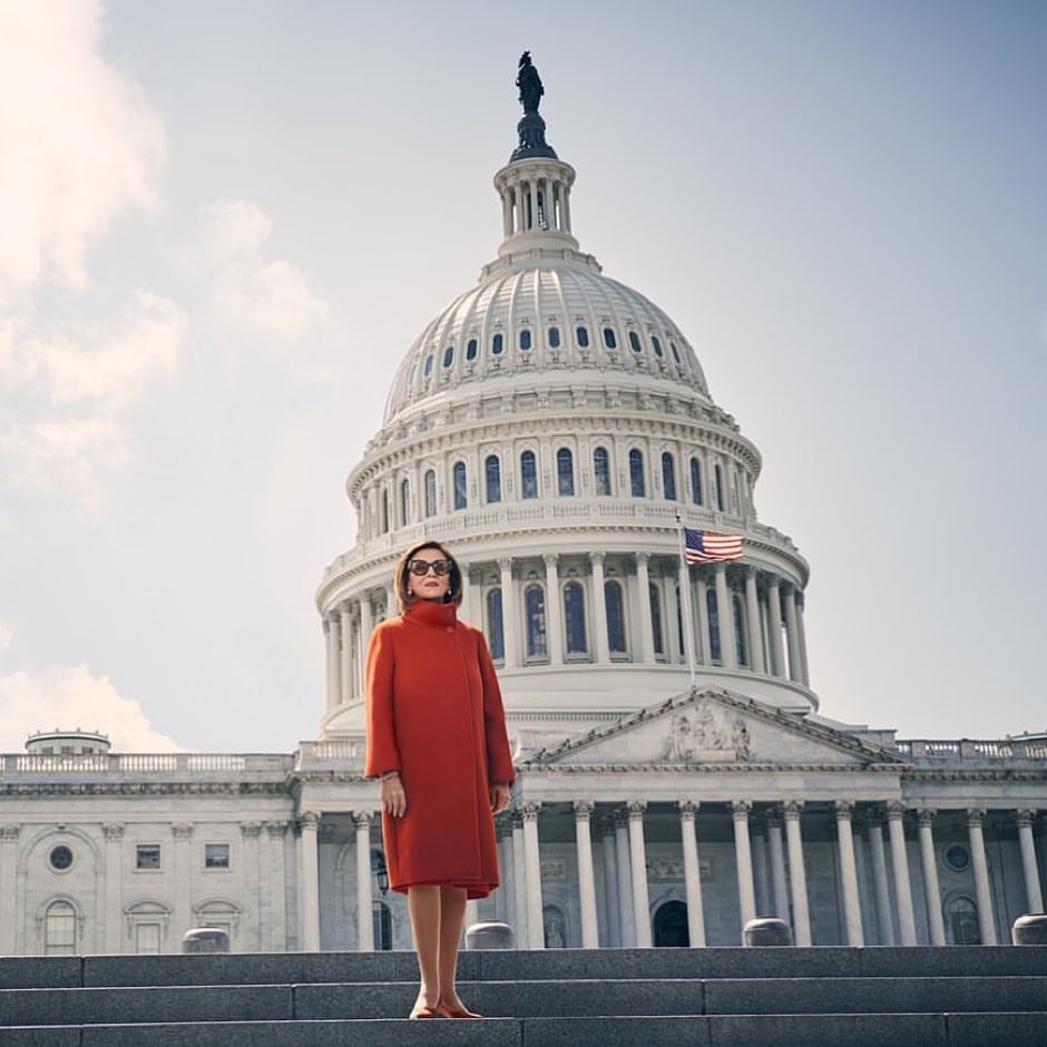 Woman in an orange coat stands on the steps of the United States Capitol Building under a sunny sky in an eye-level outdoor shot.