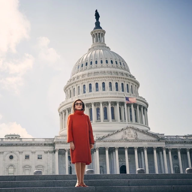 Woman in an orange coat stands on the steps of the United States Capitol Building under a sunny sky in an eye-level outdoor shot.