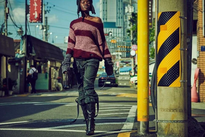 A young Black woman in a striped sweater and black boots confidently walks down an urban street with buildings and utility poles.