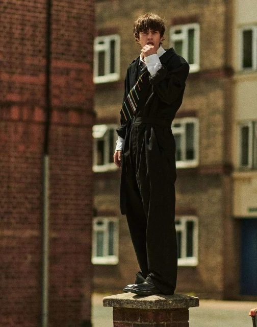 Young adult in retro-style black suit stands on a brick pillar against the backdrop of an urban apartment building on a sunny day.