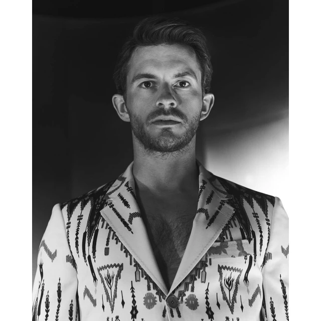 Monochrome portrait of a stylish adult male wearing an intricately patterned blazer, captured in a softly lit, modern setting.