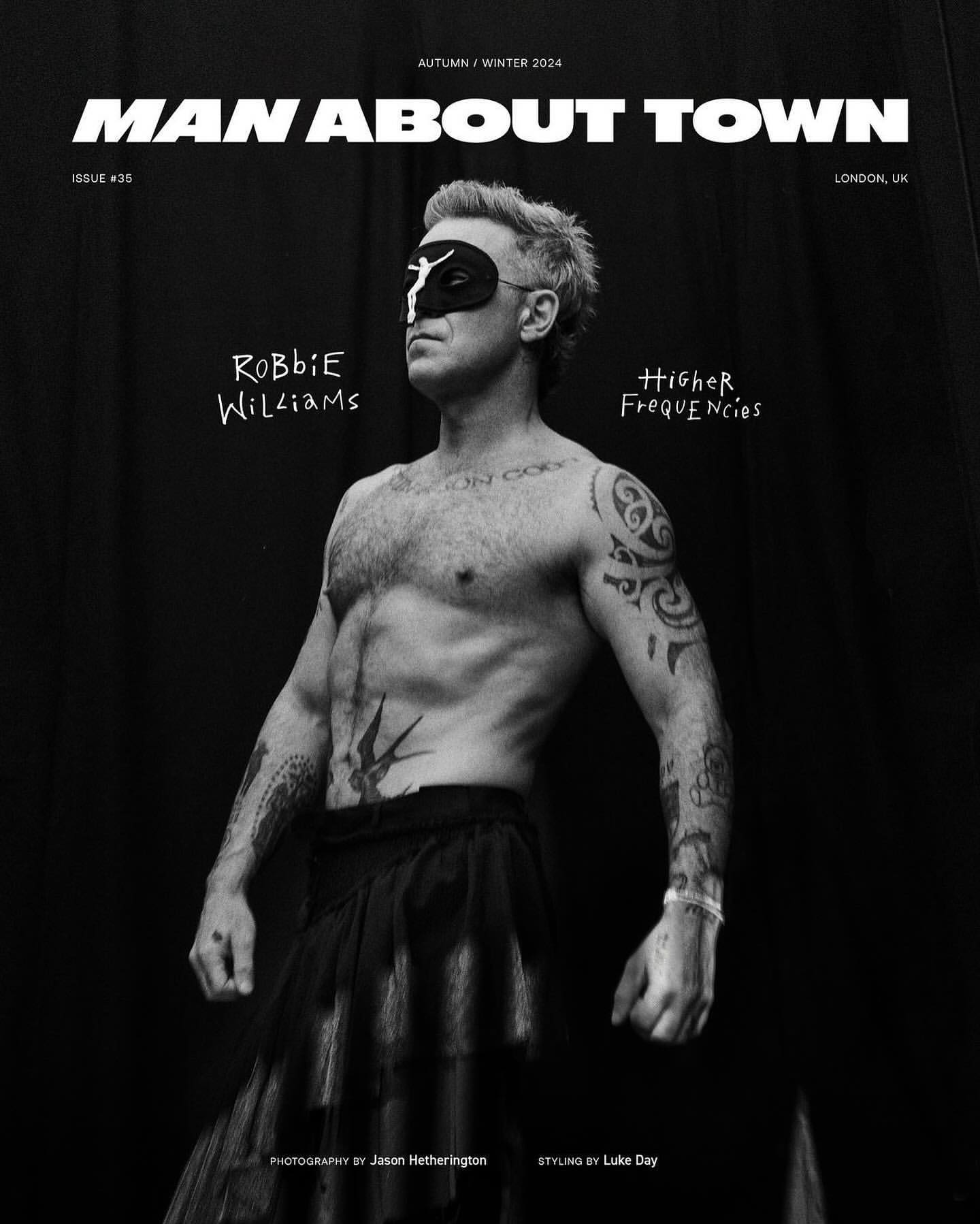 Black and white magazine cover featuring Robbie Williams wearing a mask, set against a dark backdrop with magazine text.