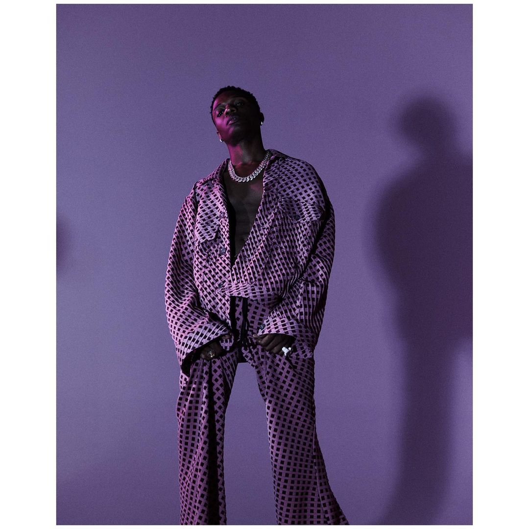 Wizkid posing in a patterned suit and jewelry, lit with violet light against a solid lilac background in a studio portrait.