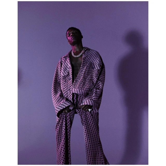 Wizkid posing in a patterned suit and jewelry, lit with violet light against a solid lilac background in a studio portrait.