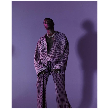Wizkid posing in a patterned suit and jewelry, lit with violet light against a solid lilac background in a studio portrait.