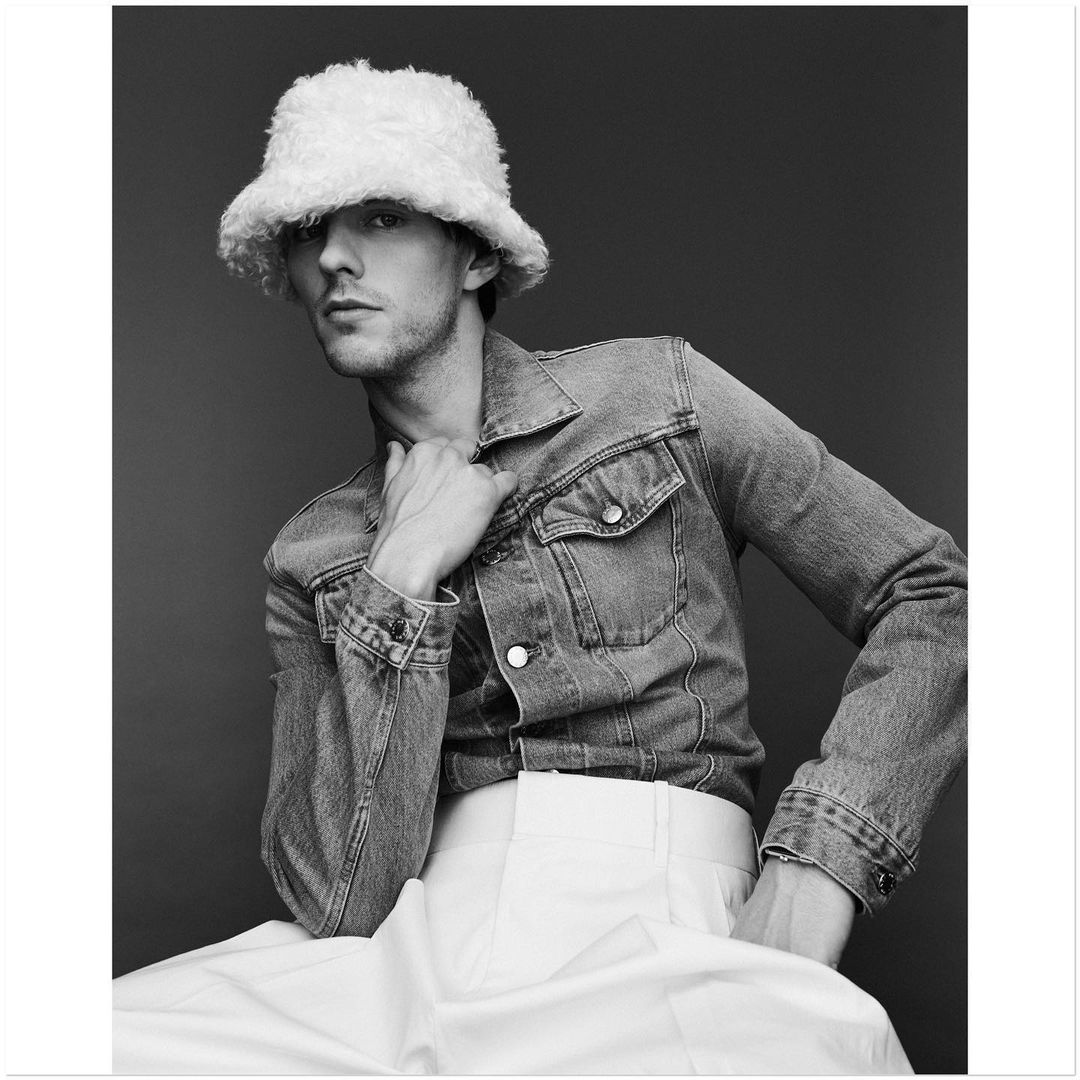 Black and white portrait of a young adult male wearing a denim jacket and a fuzzy white hat, sitting against a dark background.