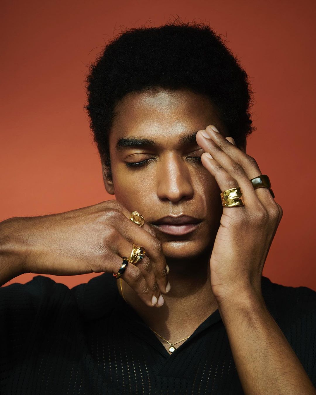 Portrait of a stylish black man with closed eyes, golden rings, and a black shirt against an orange backdrop, showcasing beauty and fashion.