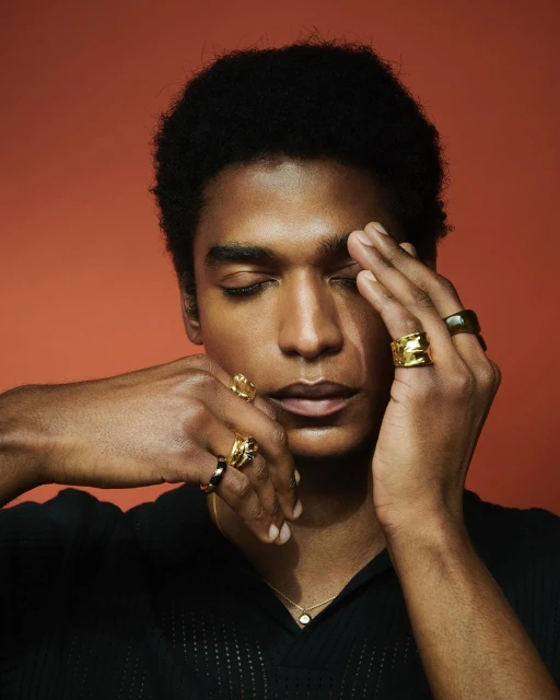 Portrait of a stylish black man with closed eyes, golden rings, and a black shirt against an orange backdrop, showcasing beauty and fashion.