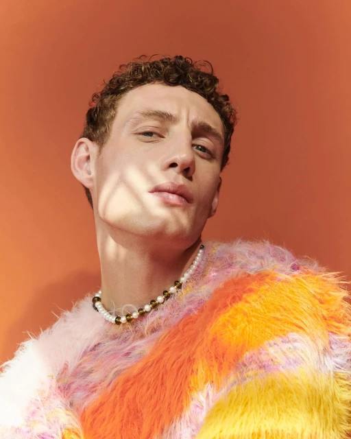 Close-up studio shot of a young adult male with curly hair, wearing a colorful sweater and beaded necklace against a orange background.