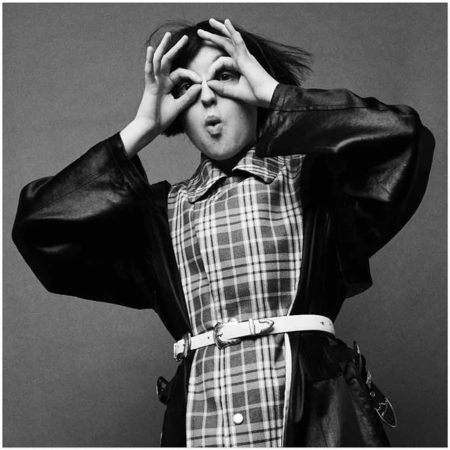 Portrait of a young person in a black jacket and plaid shirt making a funny face with their fingers posed as glasses in a studio setting.