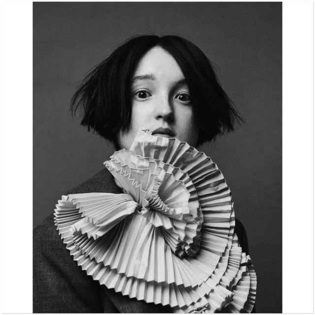 Portrait of a young woman with short dark hair wearing a ruffled collar and grey blazer in a close-up studio shot.
