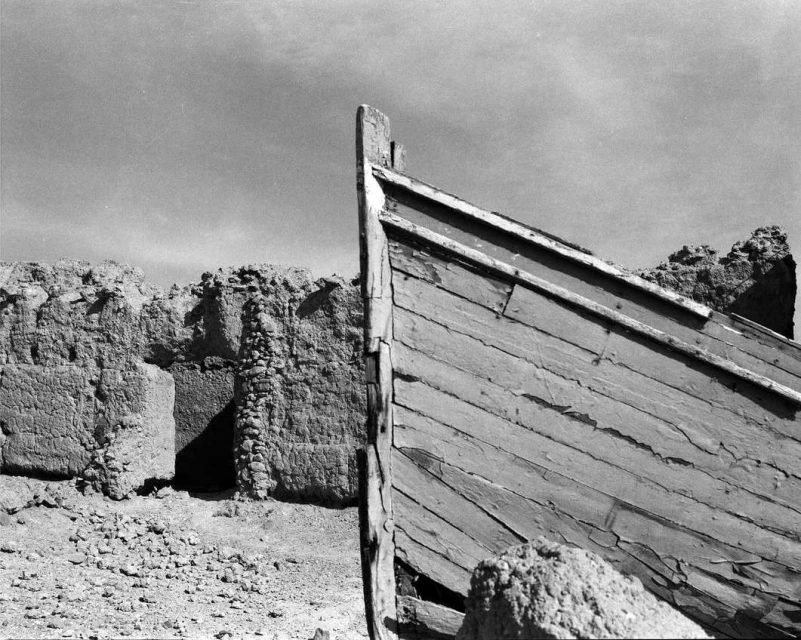 Black and white photograph of the bow of a weathered wooden boat near a crumbling, stone structure against a light sky.