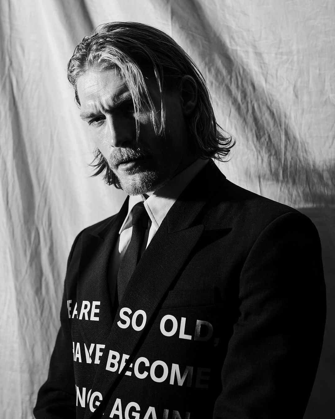 Portrait of an adult male wearing a black suit with white text, shot in black and white against a draped fabric backdrop, showcasing editorial fashion.