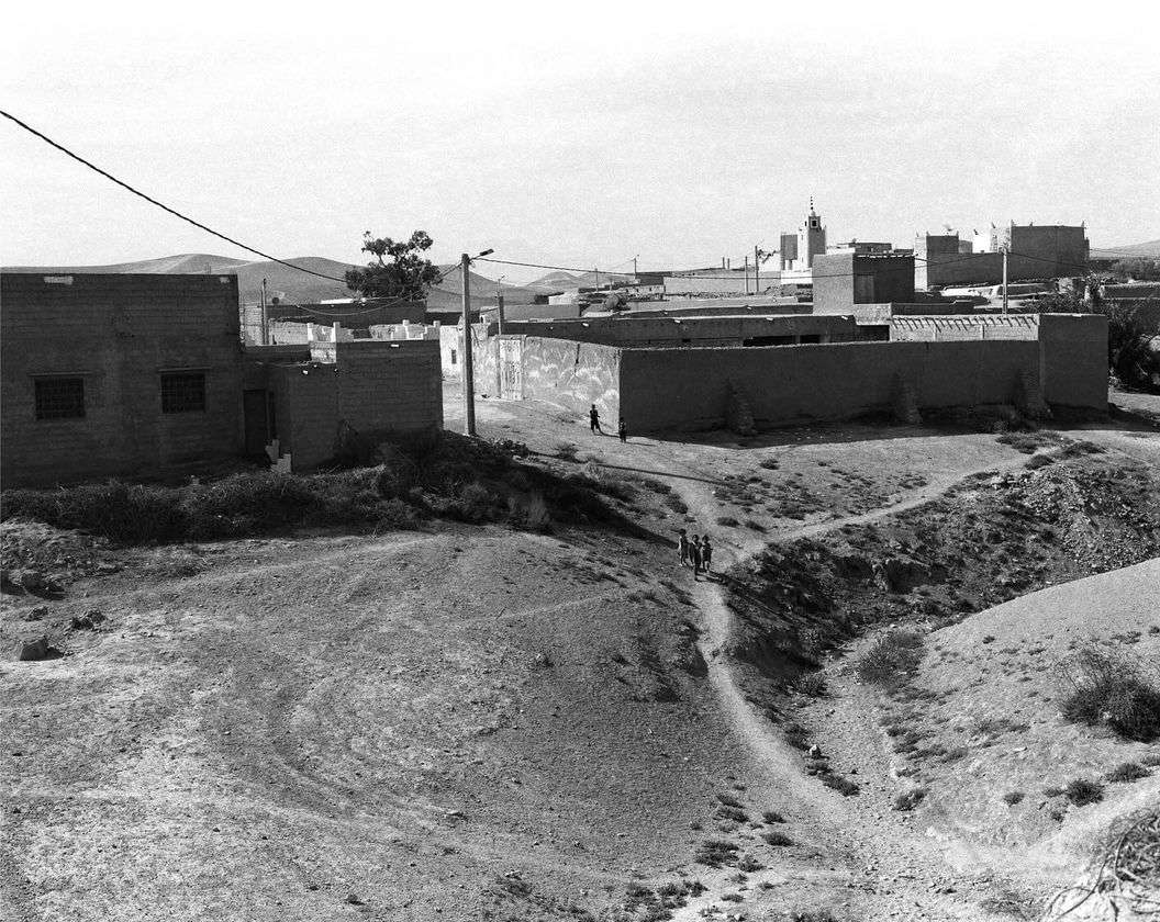 Black and white landscape view of a Middle Eastern village with adobe buildings and a group of children walking along a dirt path.