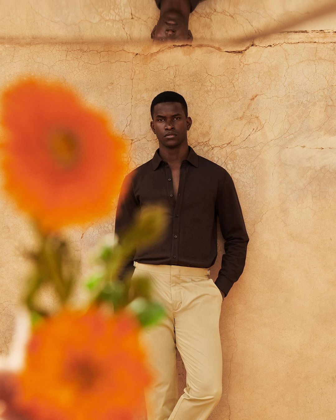Portrait of a Black man wearing a brown shirt and khaki pants standing against a textured wall with orange flowers in the foreground.