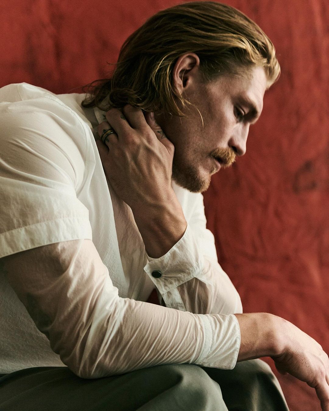 Portrait of a blonde male model in a white shirt against a reddish-brown backdrop, hand near neck, showcasing fashion and style.