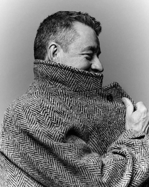 Black and white portrait of a mature man wearing a patterned tweed coat, looking away with a slight smile, indoor setting.