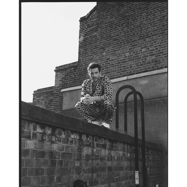 Black and white portrait of a man in a floral outfit crouching on a rooftop in an urban setting, with a brick building backdrop.