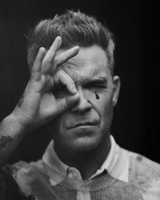 Black and white portrait of a man with hand gesture, with what appears to be a tear running down his cheek. Somber expression.