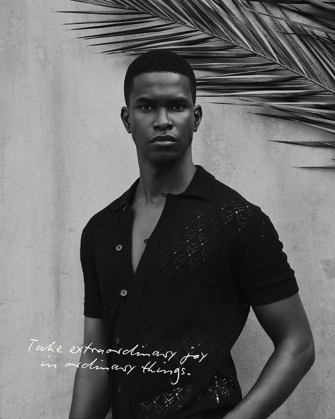 Portrait of a black male model in a black short-sleeved shirt with collar, in front of a textured gray wall with palm leaves.