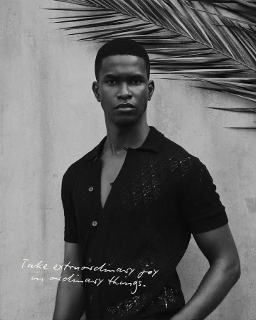 Portrait of a black male model in a black short-sleeved shirt with collar, in front of a textured gray wall with palm leaves.