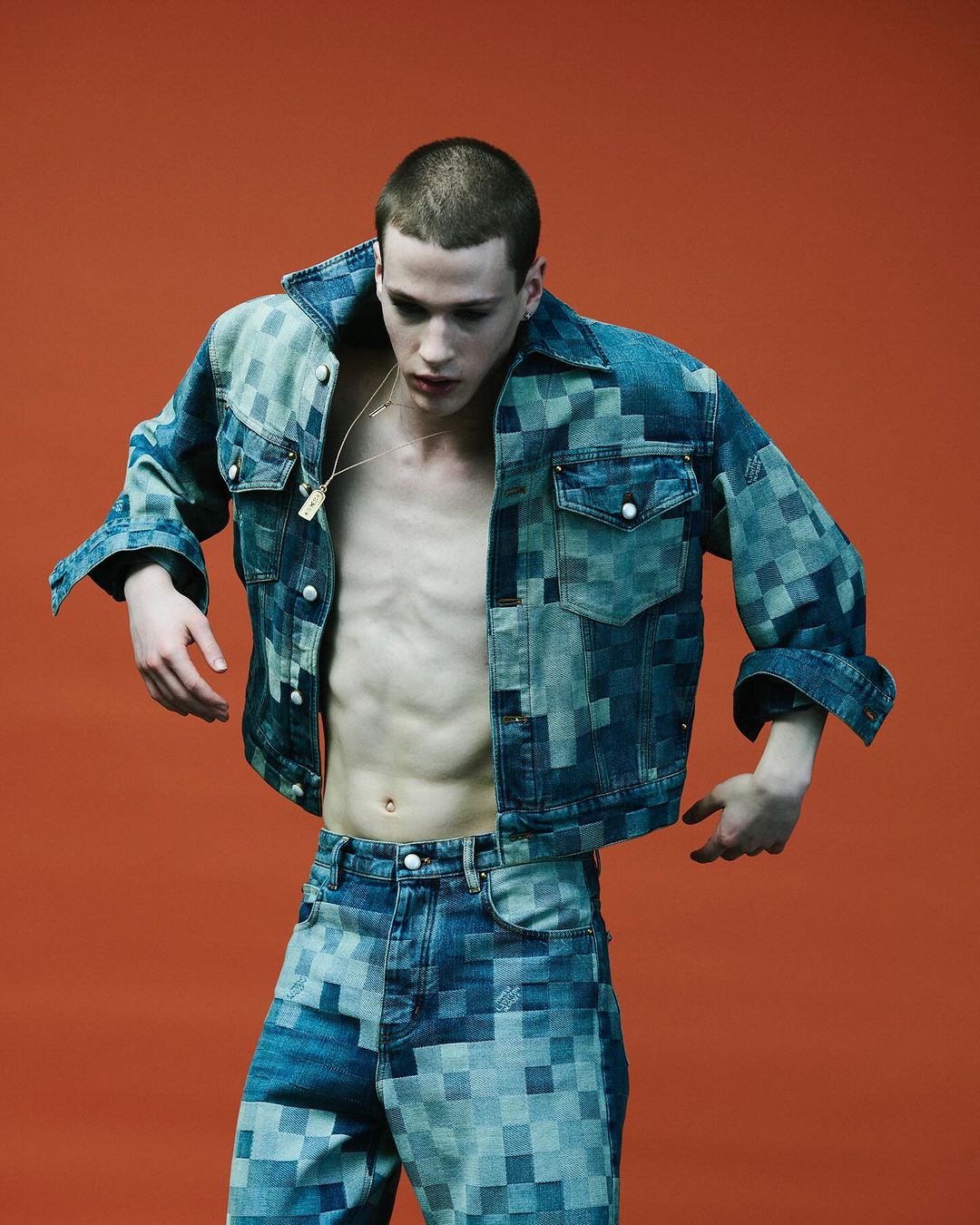 A young, slim male model poses indoors against an orange backdrop, showcasing a pixelated denim jacket and matching jeans in a fashion editorial.