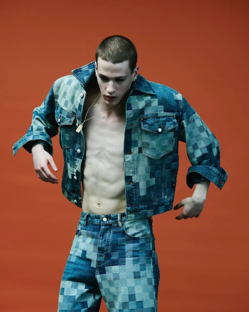 A young, slim male model poses indoors against an orange backdrop, showcasing a pixelated denim jacket and matching jeans in a fashion editorial.