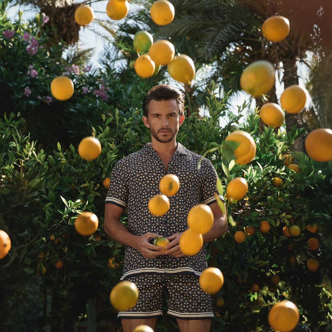 A man in a patterned shirt and shorts holds an orange, surrounded by more oranges and lush green foliage in an outdoor setting.