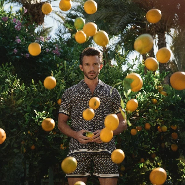 A man in a patterned shirt and shorts holds an orange, surrounded by more oranges and lush green foliage in an outdoor setting.