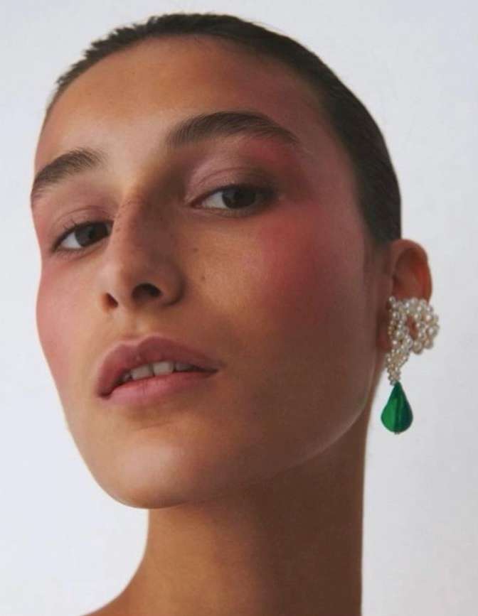 Close-up portrait of a young woman with a pearl and green gemstone earring, pink makeup, and a neutral expression against a white background.