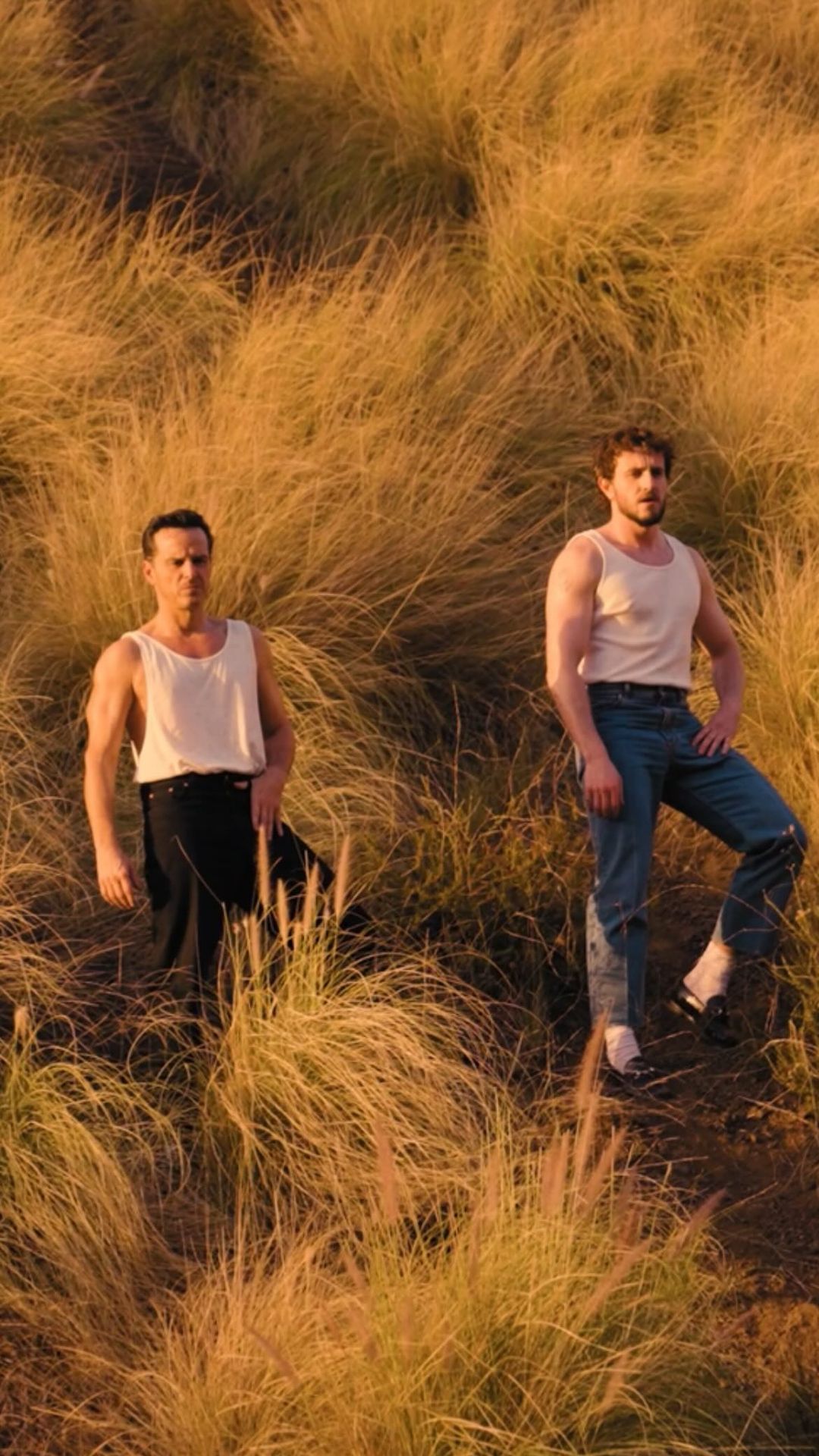 Two adult men pose in a sunlit field of tall golden grass, both wearing white tank tops and jeans, looking directly at the camera.