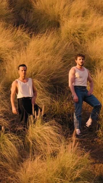 Two adult men pose in a sunlit field of tall golden grass, both wearing white tank tops and jeans, looking directly at the camera.