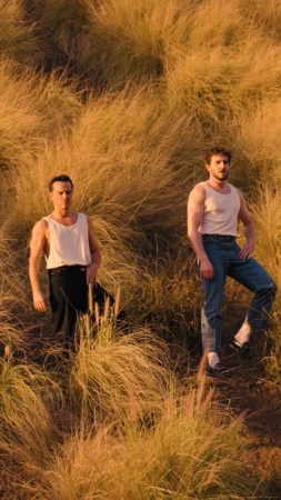 Two adult men pose in a sunlit field of tall golden grass, both wearing white tank tops and jeans, looking directly at the camera.
