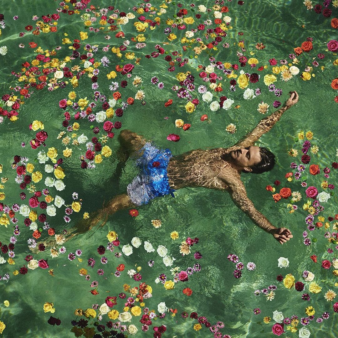 A man floats in green-tinted water among scattered flowers, viewed from directly above in this serene aquatic scene.